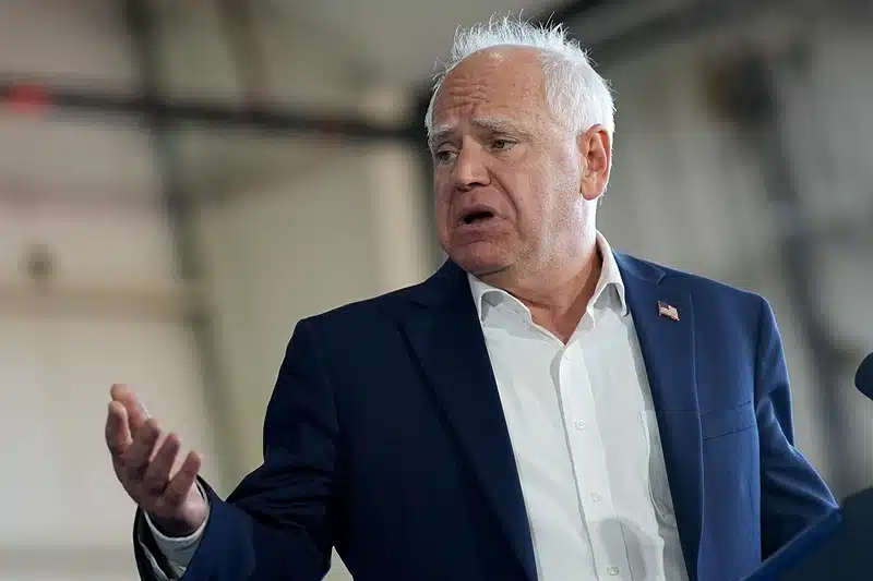 Governor of Minnesota Tim Walz in the main frame in a blue suite giving a speech. He is speaking with hand gestures and the background is blurry.