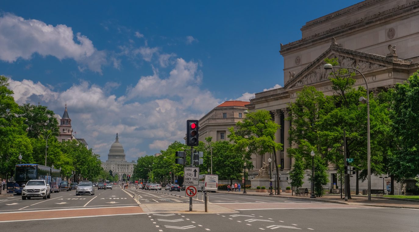 Landscape image of the capital building from far away taken from a street view. Cars, city buildings and street signs are in frame as well.